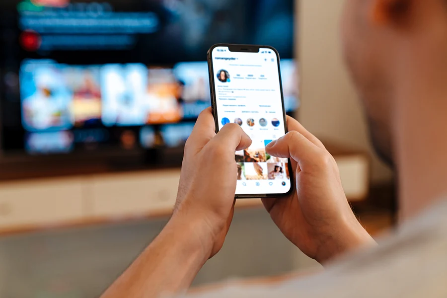 a person browsing social media on a smartphone while a streaming tv ad plays in the background, representing engagement produced by a streaming tv ads company