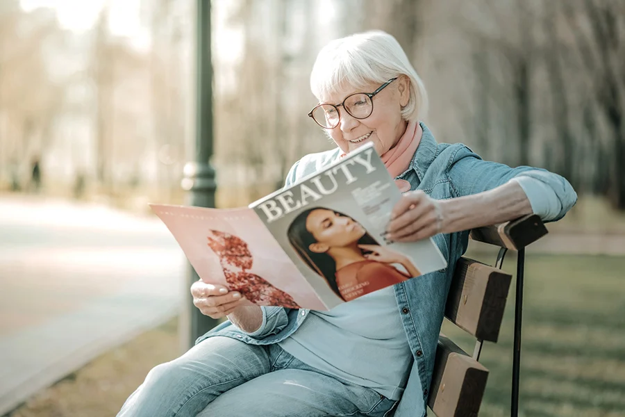 elderly woman with glasses sitting on a park bench, smiling while reading a beauty magazine highlighting the importance of advertising placement