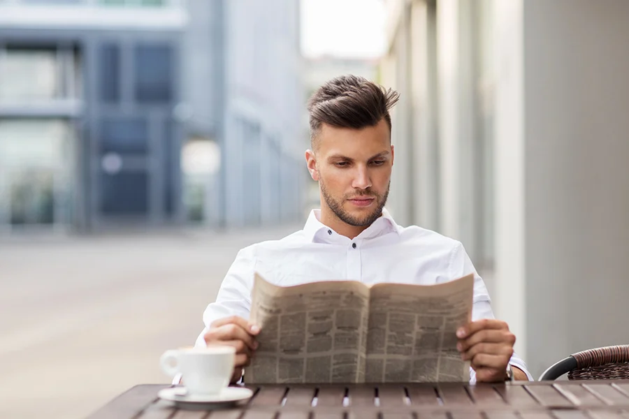 man in a white shirt reading a newspaper at an outdoor café, representing the influence of local newspaper marketing in daily life