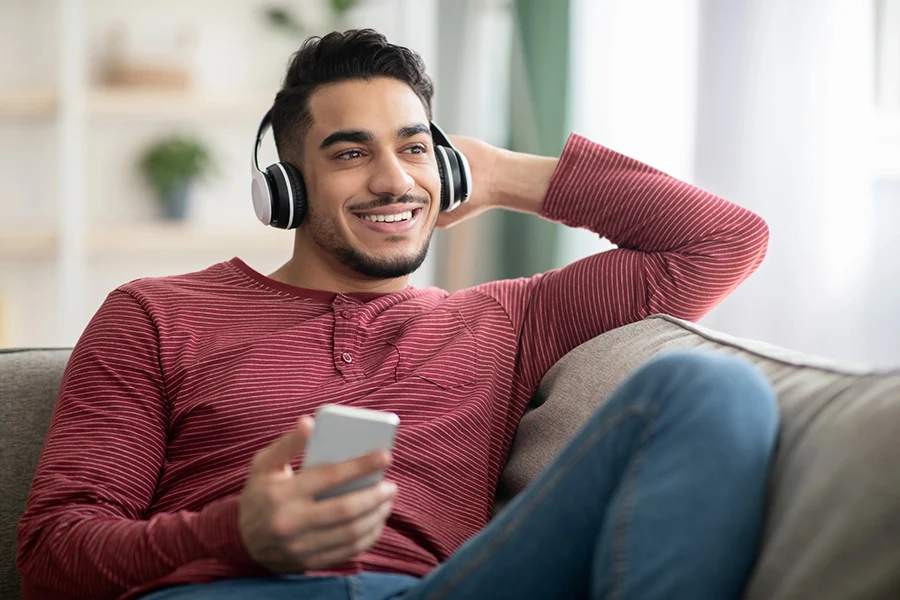 smiling man relaxing on a couch, wearing headphones and holding a smartphone, representing streaming audio advertising engagement