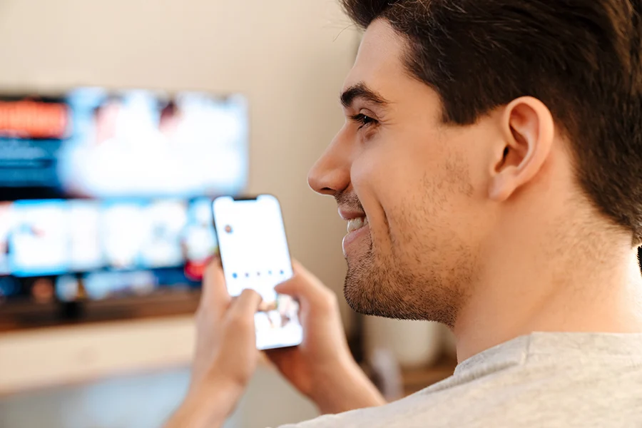 Smiling Man Using A Smartphone While Watching Tv, Illustrating Modern Viewer Behavior And Its Impact On Advertising Rates For Cable Tv smiling man using a smartphone while watching tv, illustrating modern viewer behavior and its impact on advertising rates for cable tv