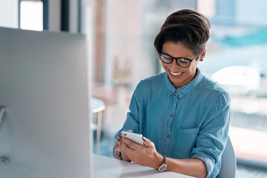 smiling woman in glasses using a smartphone at her desk, engaging with network broadcasting advertising content
