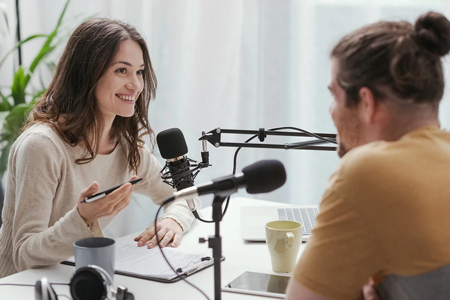 smiling woman interviewing a guest in a professional podcast setup with microphones, headphones, and a laptop, illustrating the power of podcast advertising