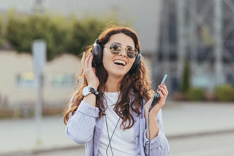 Smiling Woman Wearing Headphones And Holding A Smartphone, Enjoying Music Streaming Via Satellite Radio Outdoors smiling woman wearing headphones and holding a smartphone, enjoying music streaming via satellite radio outdoors