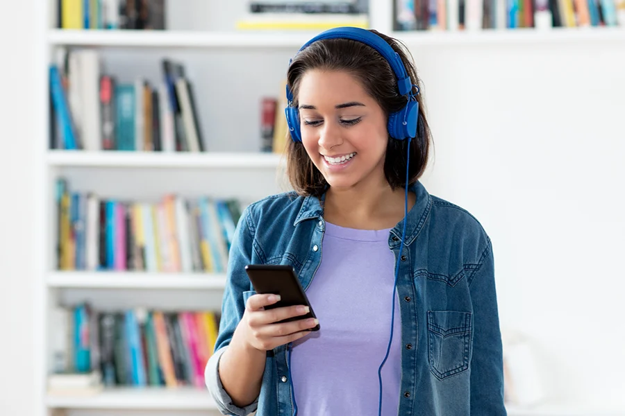 smiling young woman wearing blue headphones, listening to a podcast on her smartphone, highlighting the impact of podcast audio advertising
