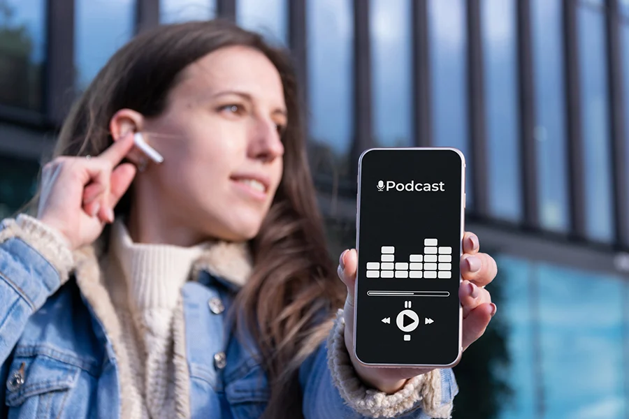 oung woman outdoors inserting wireless earbud while holding smartphone displaying a podcast app, showcasing digital audio marketing trends