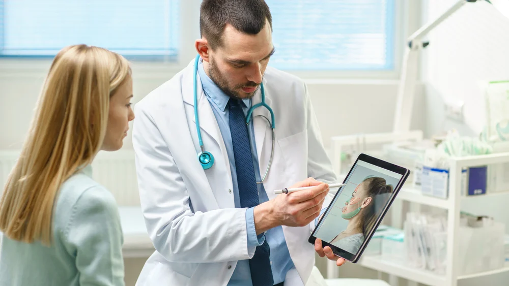 Plastic Cosmetic Surgeon Consults Woman About Facial Lift Surgery, He Draws Arrows On Digital Tablet Computer Screen, Showing Types Of Facelift, Forehead Correcting Procedures Available For Her. plastic surgeon consulting with patient